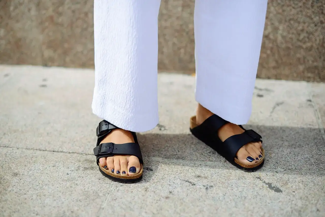 Pies con sandalias Birkenstock negras, ideales para aliviar la fascitis plantar. Pantalones blancos y uñas oscuras.