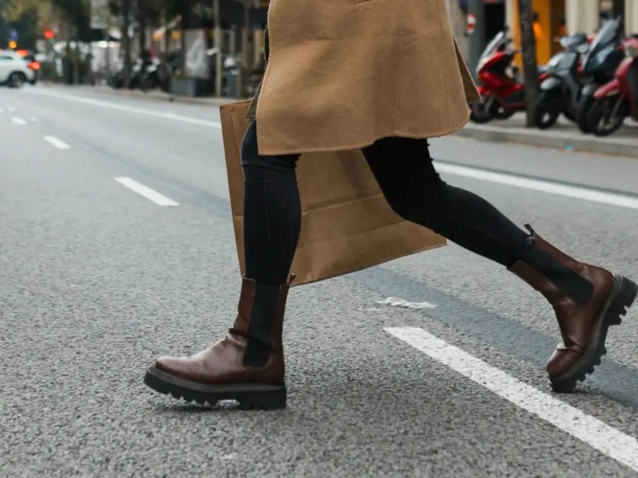 Mujer con botas chelsea marrones y abrigo camel cruza la calle, llevando una bolsa de compras. Un outfit perfecto para el día a día.
