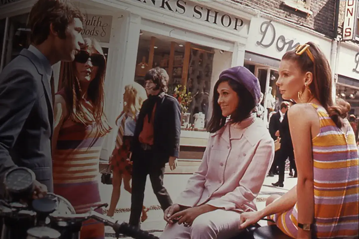 Dos mujeres con estilo mod, una con boina morada y chaqueta rosa, la otra con vestido a rayas, pasean por una calle comercial.