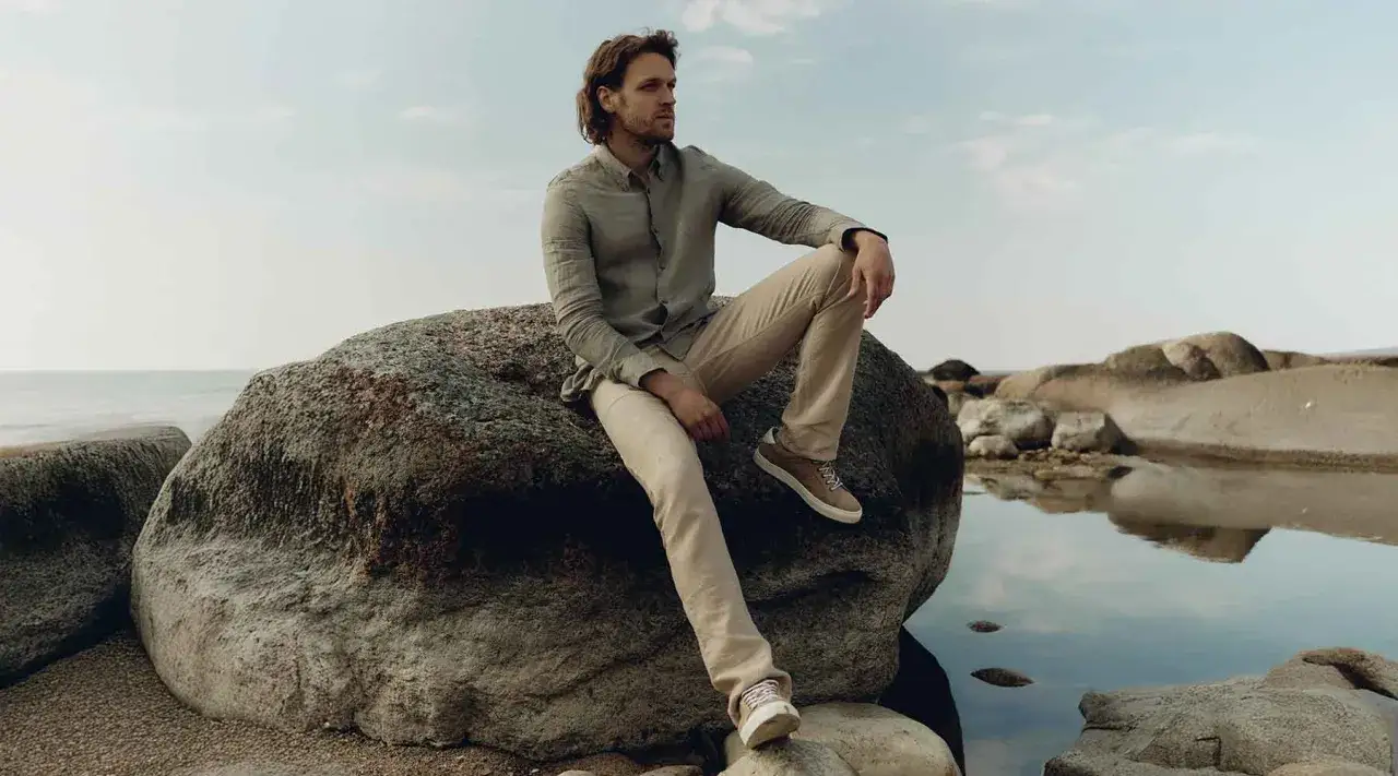 Hombre con camisa y pantalón lino beige, sentado en roca junto al mar.