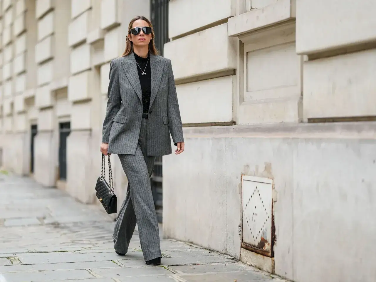 Mujer con un elegante power suit gris a rayas, gafas de sol y bolso negro, caminando por la calle.