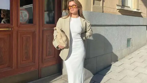 Mujer con blazer y vestido largo blanco, bolso tejido y gafas de sol, posando frente a una puerta de madera.